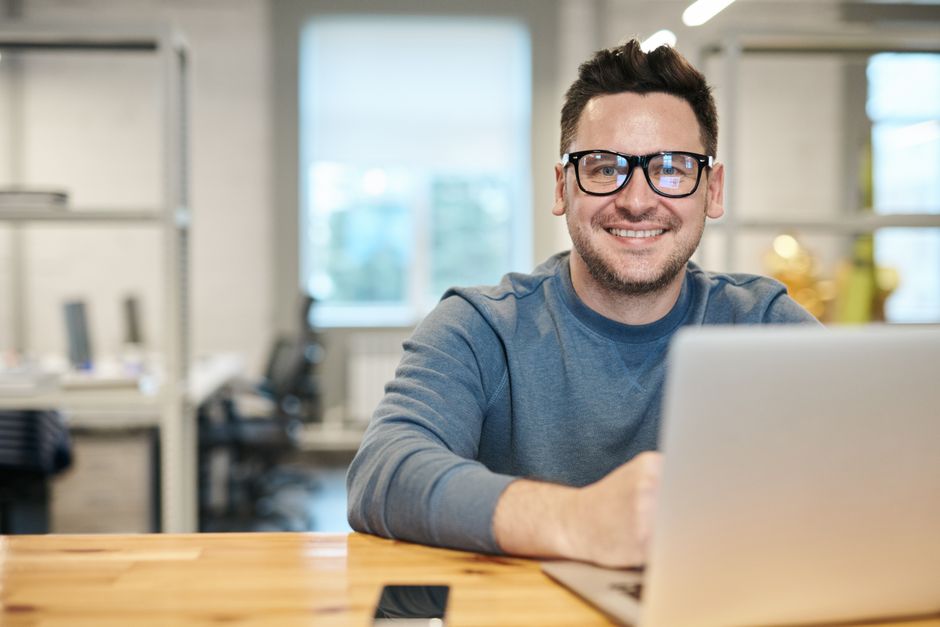 A man in glasses working at a laptop in an office, looking up and smiling.