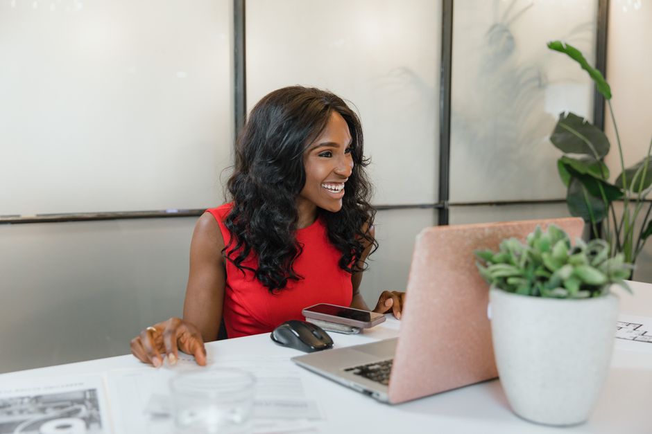 A woman smiling while working at a laptop with a phone beside her at a bright desk.